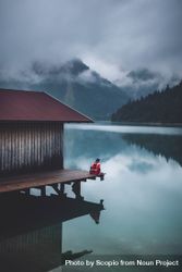 Person sitting on dock over lake under cloudy sky 0WAgr4