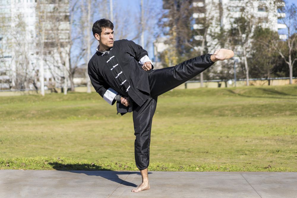 Young athletic martial arts fighter practicing kicks in a public park wearing classic kung fu uniform