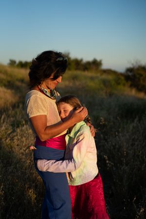 Woman hugging her daughter looking down and smiling outside in canyon at sunset