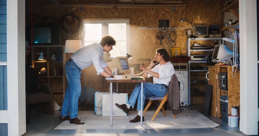 Two White Male Stock Brokers Working In Retro Garage, Using Old Desktop Computer With Candlestick Chart And Landline Phone To Pitch A Public Company To Investor.