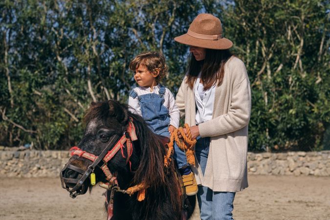 Smiling young woman standing near adorable toddler riding horse