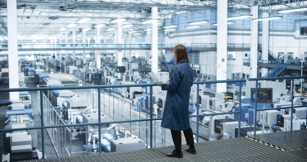 Japanese Factory Manager Working on Laptop at a Futuristic Industrial Manufacture with Autonomous AI Robots. Specialist Monitoring All the Data on a Computer, Checking Reports and Planning Research