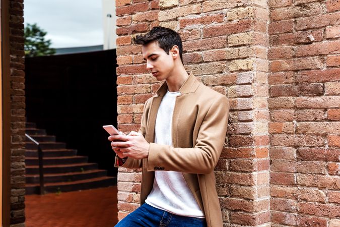 Man in camel coat texting on phone while leaning on brick wall