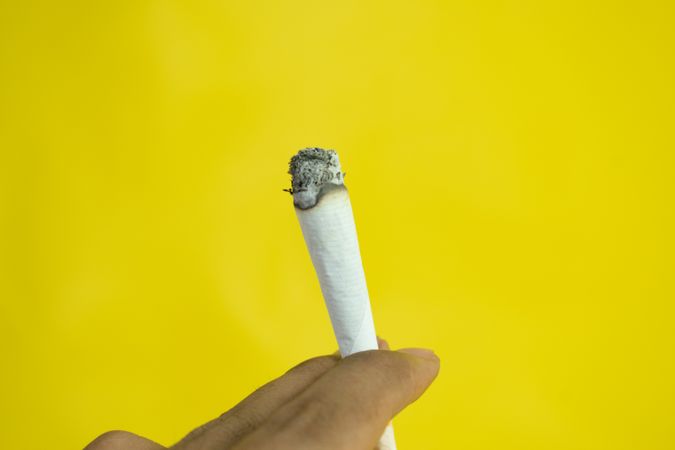Fingers holding burning hand rolled cigarette on yellow background