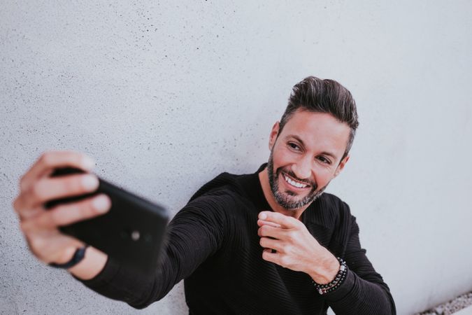 Adult stylish smiling man taking selfie on smartphone near grey wall