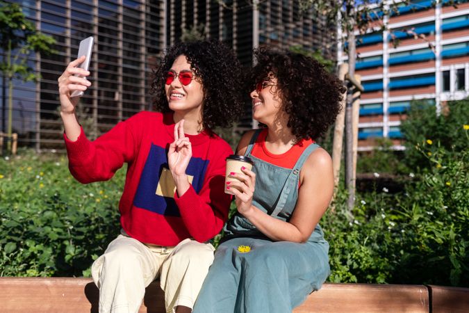 Two female friends sitting on bench outside taking selfie on phone together