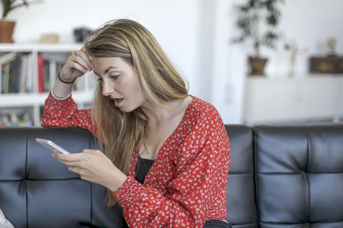 Surprised young blond woman using mobile phone app on sofa