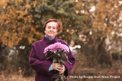Older woman in purple coat holding flower bouquet standing near yellow trees bxKpr4