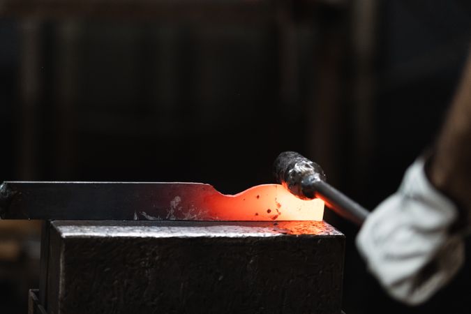 Blacksmith in gloves hot hitting metal part on anvil