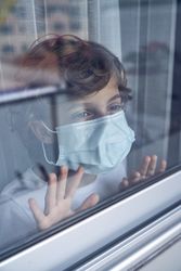Pensive boy in mask standing alone and looking out of window at