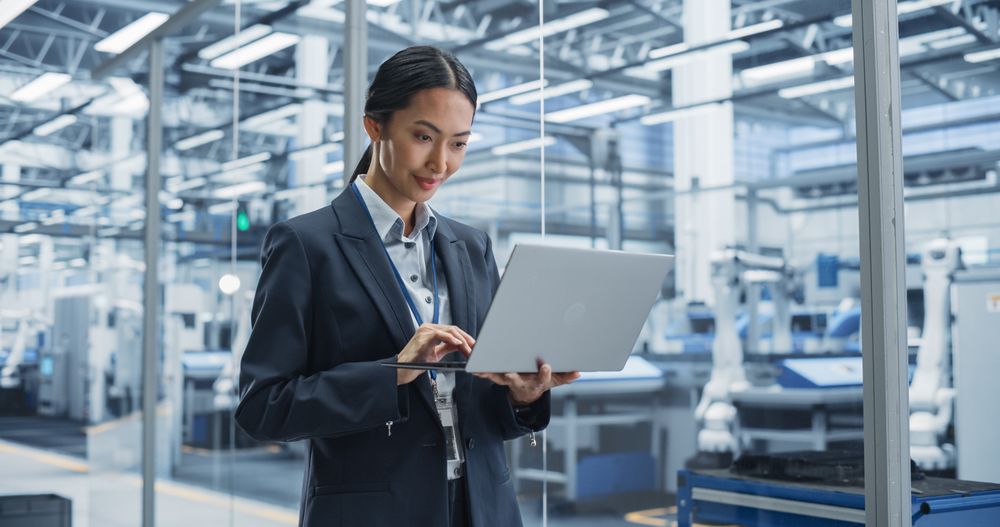 Asian Factory Manager Working on Laptop at a Futuristic Industrial Manufacture with Autonomous Robotic Assembly Line. Specialist Monitoring Production, Planning Work for Employees