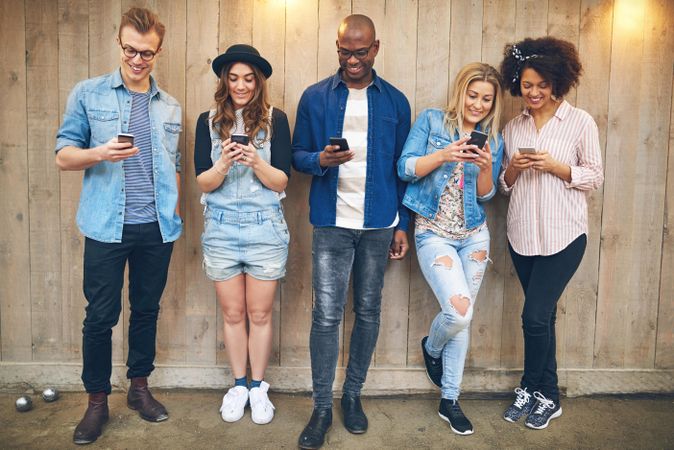 Multi-ethnic group of friends standing against a wooden fence looking down at their phones