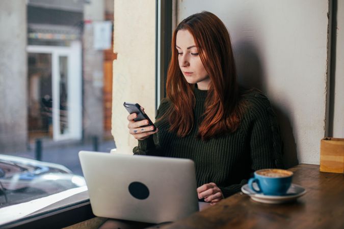 Woman checking phone in cafe with laptop