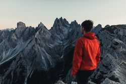 Back view of man in red jacket standing against mountainous landform in Dolomites, Italy 5XJ9Mb