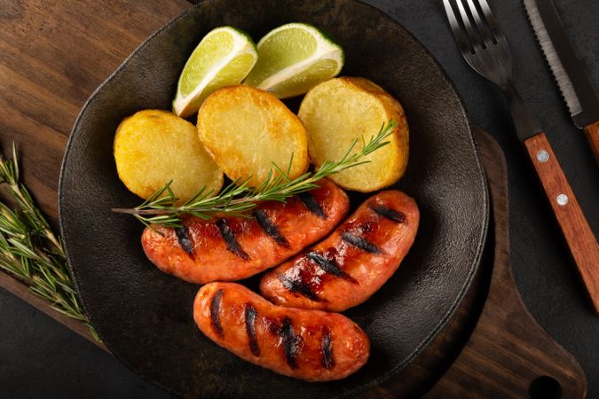 Top view of plate with grilled sausages, potatoes, lime slices and sprig of rosemary