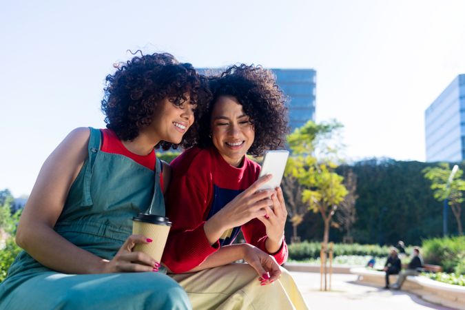 Happy female friends hanging in park together drinking coffee, checking phone