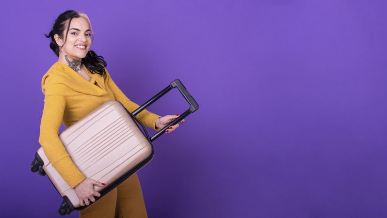 happy woman traveler holding her suitcase like a guitar against purple background. Copy space.
