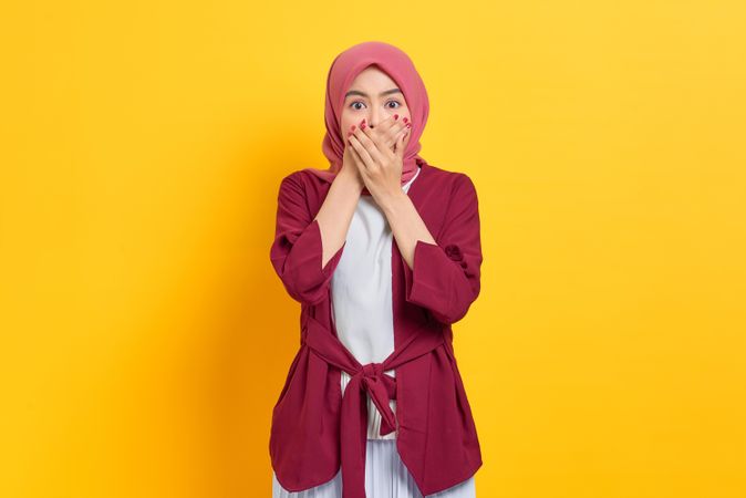 Surprised woman in red headscarf looking at camera with both hands over her mouth