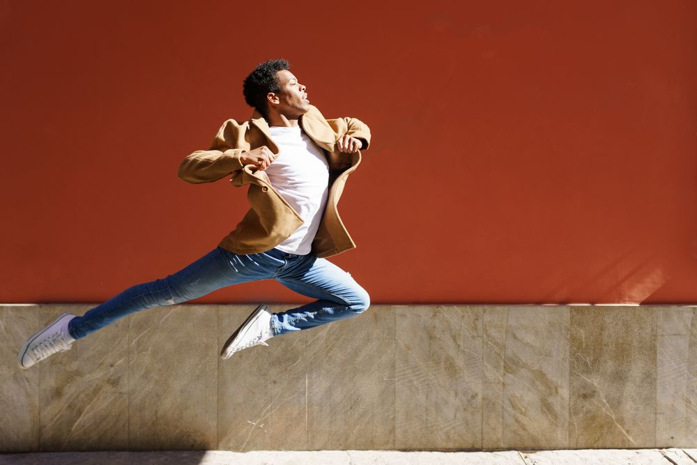Black male in camel coat leaping against am outdoor wall