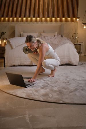 Woman with rolled yoga mat using laptop in bedroom