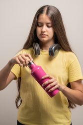 A young woman in a studio opening a reusable pink water bottle to stay hydrated.