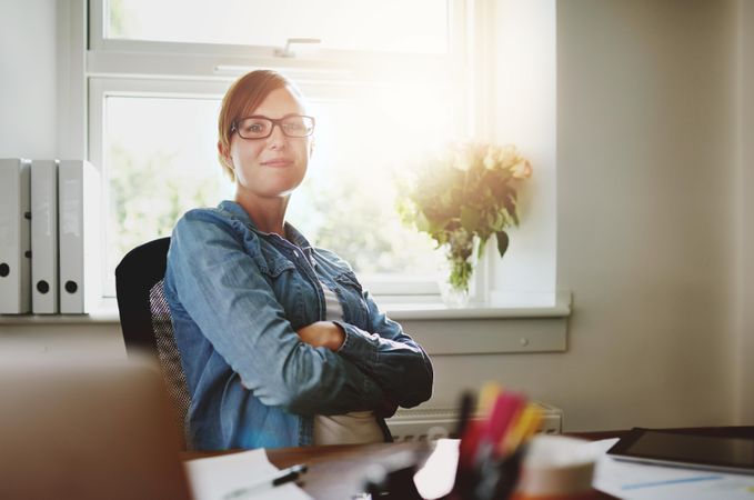 Confident female entrepreneur in jean jacket sitting at her desk in bright office