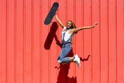 Woman smiling and jumping for joy in front of a red wall with skateboard