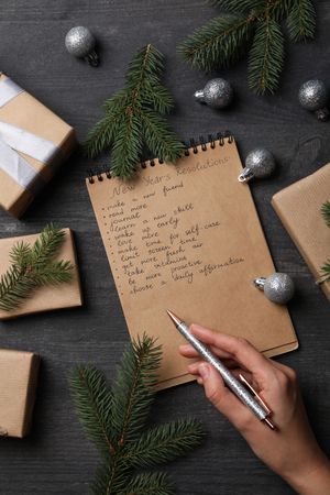 Notepad with list, gift boxes and pen in hand on gray wooden background, top view