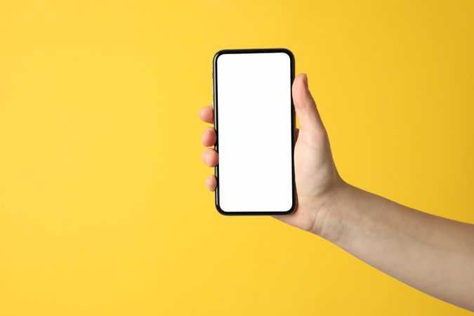 Female hand holding phone with empty screen on yellow background