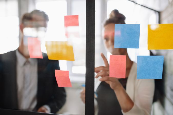 Business woman pointing at sticky note to male colleague on glass wall in office