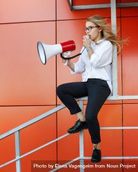 Woman in glasses sitting in front of orange wall speaking into megaphone, vertical bGaza5