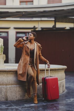 Stylish Black woman talking on smartphone with luggage on street