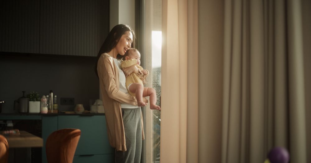 Young Asian Woman Holding her Baby in her Arms While Standing Next to a Window at Home.