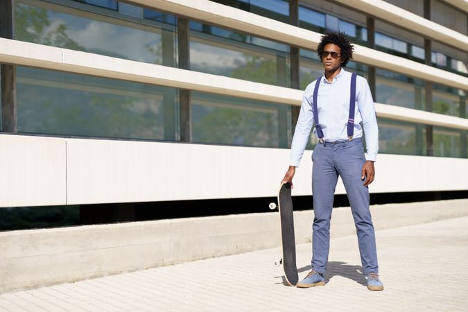 Serious man in sunglasses standing tall with skateboard outside building