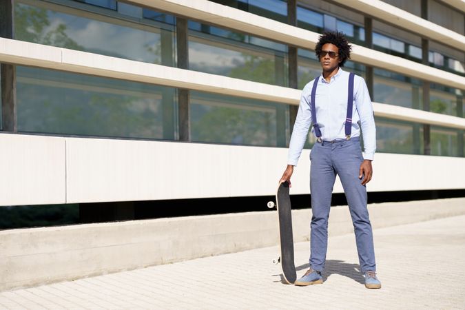 Serious man in sunglasses standing tall with skateboard outside building