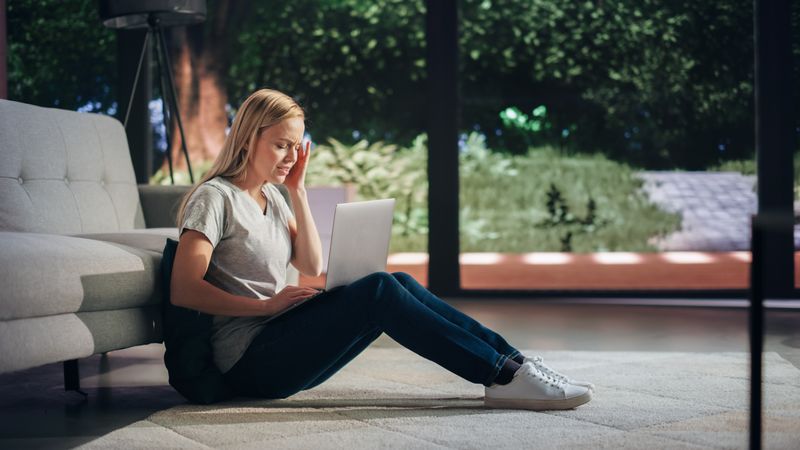 Portrait of Beautiful Young Adult Female Sitting on a Floor, Making a Video Call on Laptop Computer And Experiencing Sudden Headache.