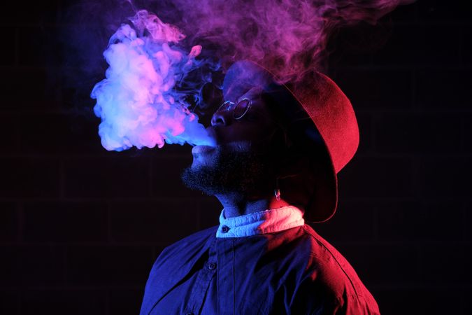 Stylish Black man in hat vaping against brick wall