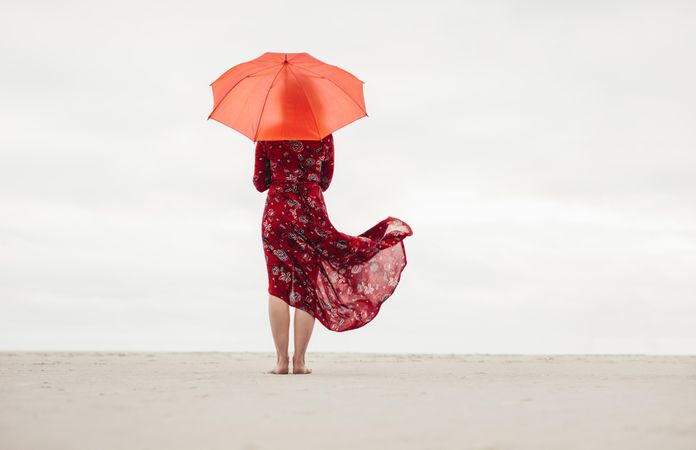 Rear view of a woman in red dress holding umbrella standing on the beach