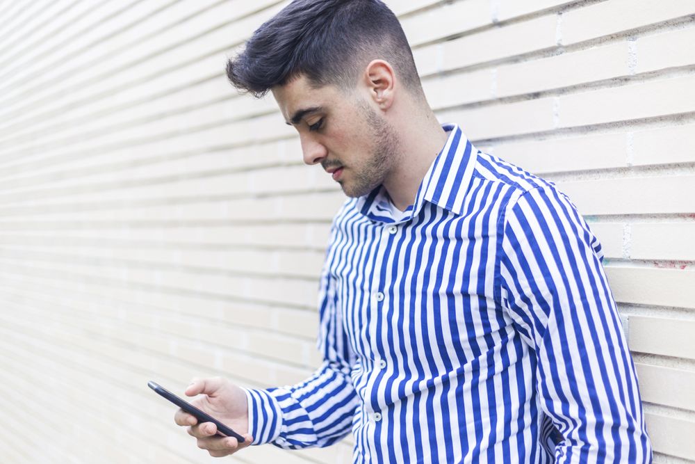 Young happy man leaning on house wall while using his smartphone