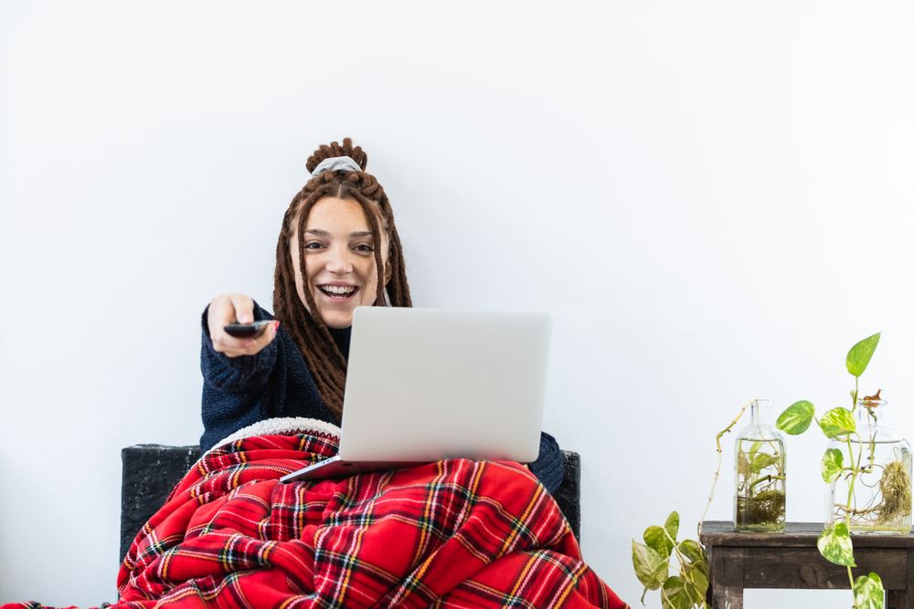 Woman with dreadlocks working with a laptop at home and with a remote control in hand