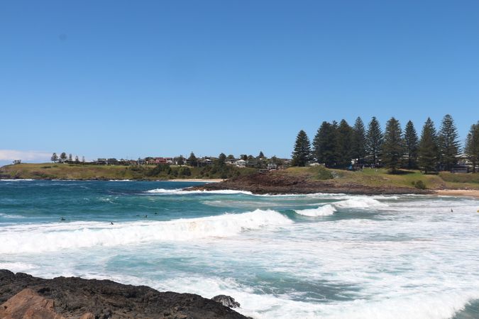 Waves crashing on rocks at a coastal area with palm trees and homes under clear blue skies