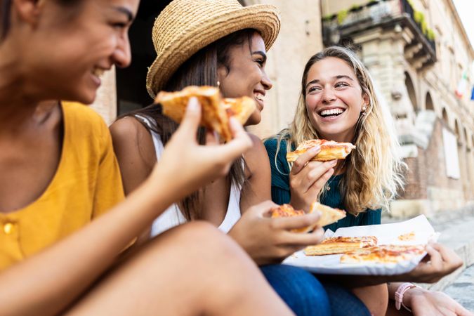 Three young multi-ethnic women laughing while eating a piece of pizza in Italian city street