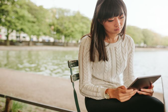 Shot of young Asian woman sitting on chair by the pond and using digital tablet