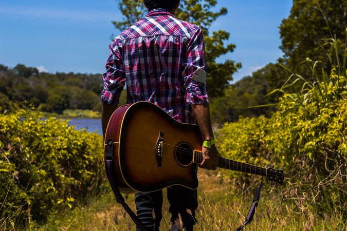 Back view of man in red and blue plaid shirt holding a guitar and walking in woods