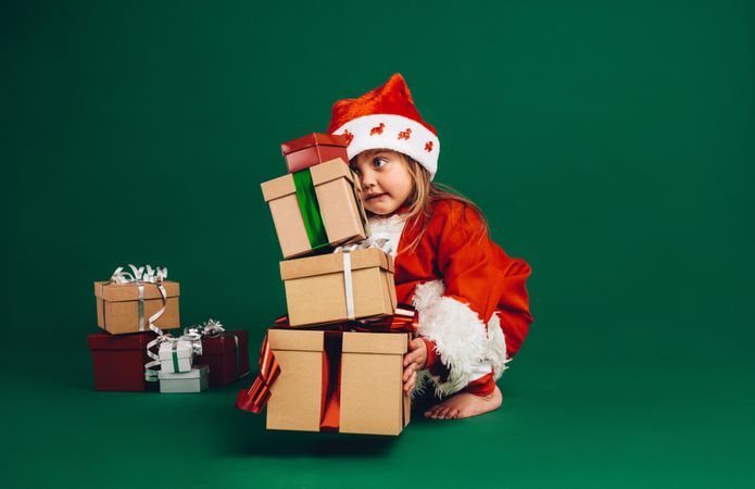Cute girl wearing festive costume and Santa hat bending down to carry gifts