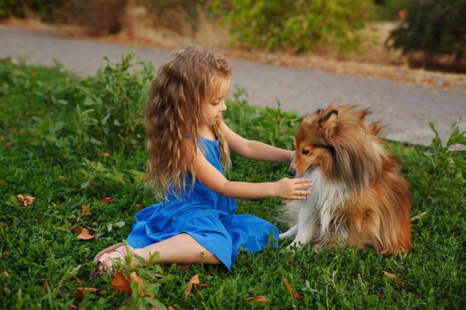 Adorable child in blue dress playing with pet dog in the grass