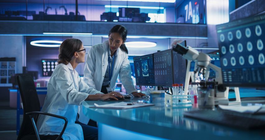 Asian And White Female Doctors Talking In Medical Research Center And Using Desktop Computer To Analyze Brain CT Scans.