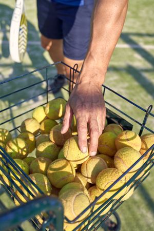 Crop man taking tennis ball from basket on playground