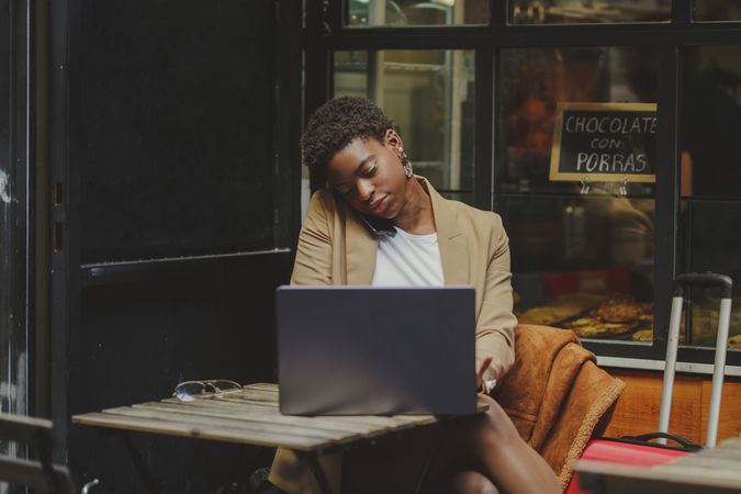 Black woman talking on smartphone and sitting at table with laptop in cafe