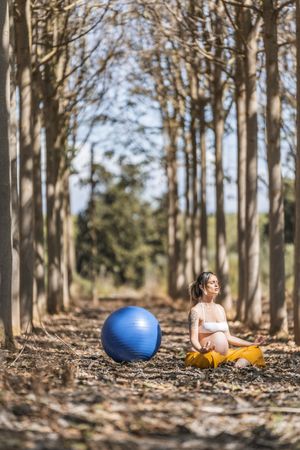 Serene adult pregnant woman meditating while sitting on ground among trees in park during sunny day